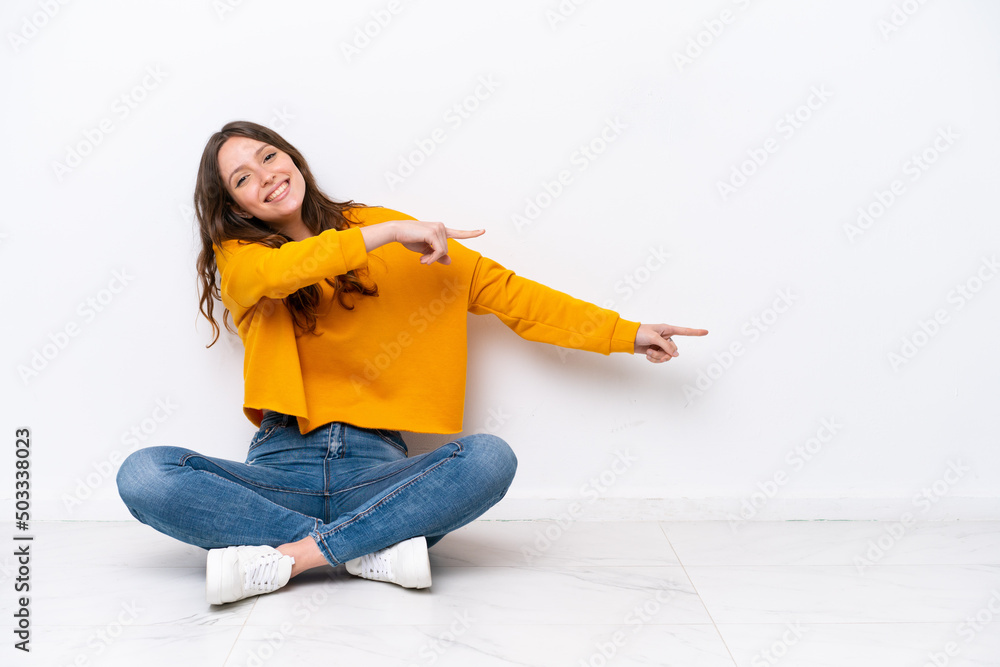 Young caucasian woman sitting on the floor isolated on white wall pointing finger to the side and presenting a product Young caucasian woman sitting on the floor isolated on white wall pointing finger to the side and presenting a product