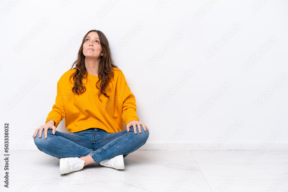 Young caucasian woman sitting on the floor isolated on white wall and looking up Young caucasian woman sitting on the floor isolated on white wall and looking up