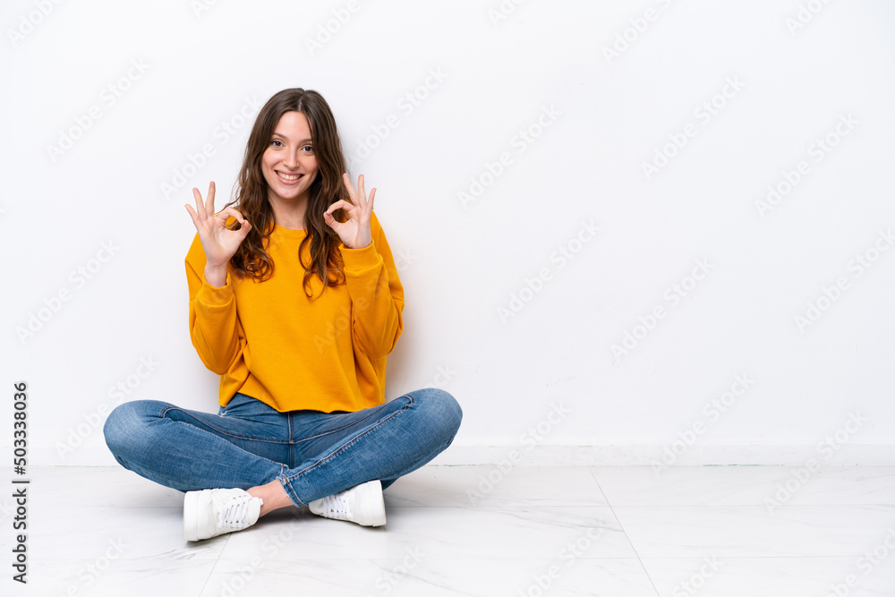 Young caucasian woman sitting on the floor isolated on white wall showing an ok sign with fingers