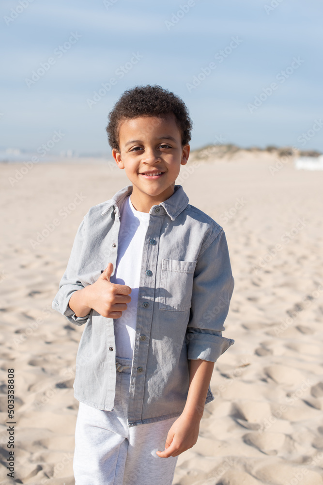 Portrait of African American boy on beach. Male model with curly hair in casual clothes looking at camera, showing thumb up. Portrait, beauty concept Portrait of African American boy on beach. Male model with curly hair in casual clothes looking at camera, showing thumb up. Portrait, beauty concept