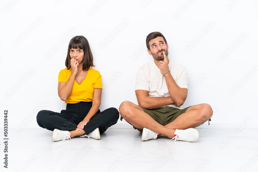 Young couple sitting on the floor isolated on white background having doubts while looking up Young couple sitting on the floor isolated on white background having doubts while looking up