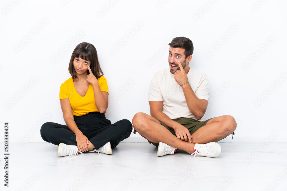 Fototapeta premium Young couple sitting on the floor isolated on white background looking to the front