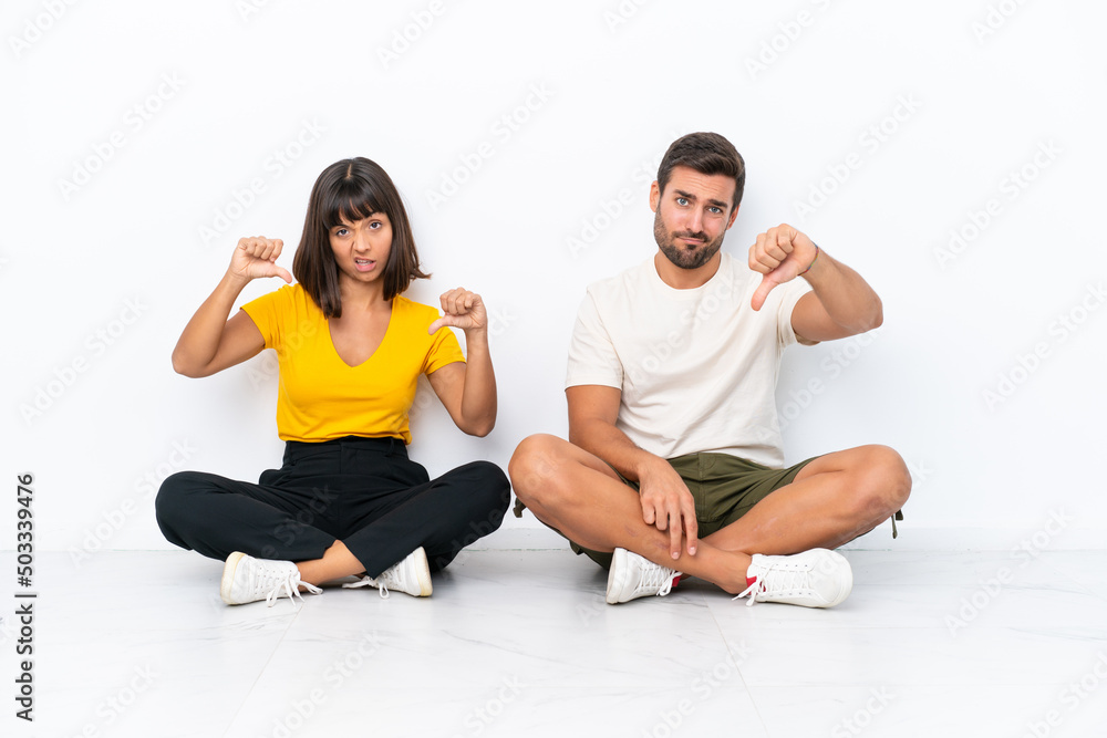 Young couple sitting on the floor isolated on white background showing thumb down with both hands Young couple sitting on the floor isolated on white background showing thumb down with both hands