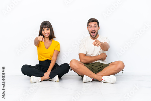 Young couple sitting on the floor isolated on white background points finger at you with a confident expression