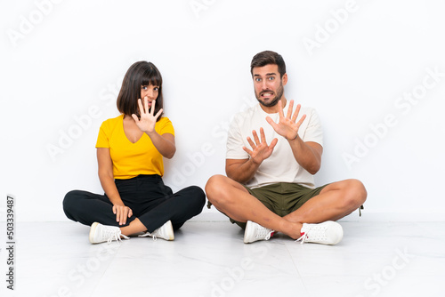 Young couple sitting on the floor isolated on white background is a little bit nervous and scared stretching hands to the front