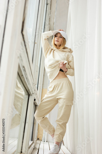 Posing fitness woman in her 20s casual sportswear tracksuit: trainers, cap, hoodie sweatshirt, pants set(jogging, gym) near the window. Female sport fashion, cozy, stylish and comfortable. Vertical