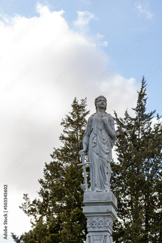 St. Thomas, Ontario, Canada - November 13 2021: Selective focus of tall gravestone statue of boy in robes with anchor by his side. Against blue sky and trees.