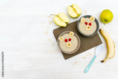 Funny cute kids childrens baby's healthy breakfast lunch oatmeal porridge in bowl look like bear face decorated with apple, banana, dried berry fruits. dessert food art on white wooden table