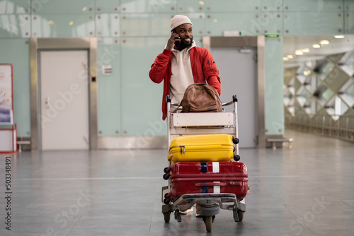 Young smiling African American man pushing luggage trolley while walking after arrival at airport, talking on mobile phone. Happy Black male tourist rolling a baggage cart in terminal. Trip, journey.