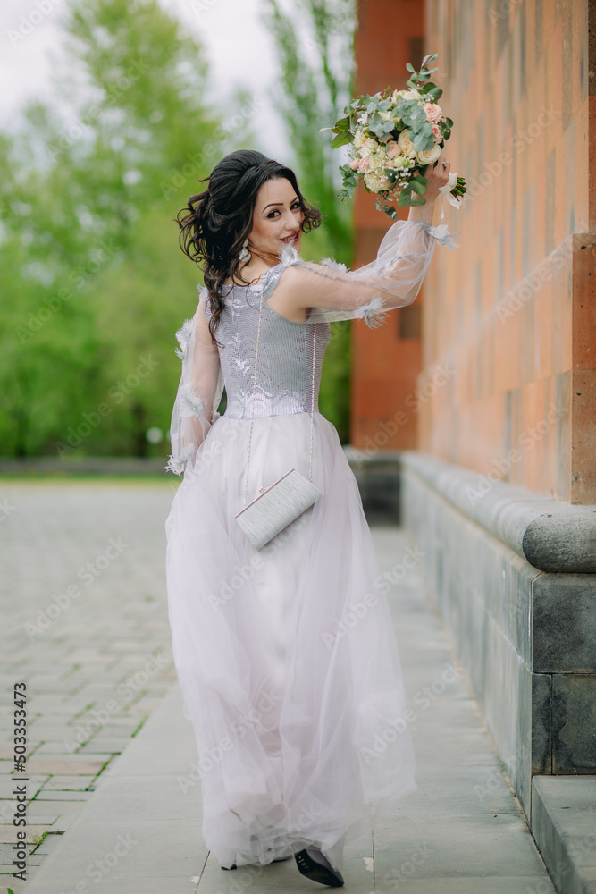Active smiling bridesmaid with elegant dress standing next to the church and showing bridal bouquet