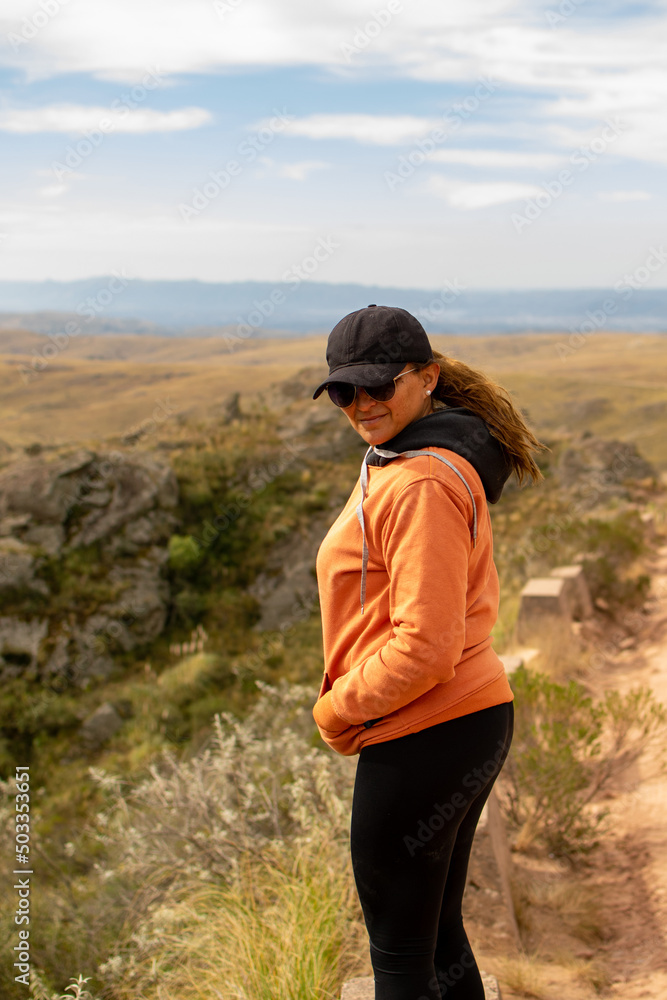 Portrait of latin woman dressed in orange with a cap and tail back having fun during the day of trekking in the mountain forest - Focus looking at the camera