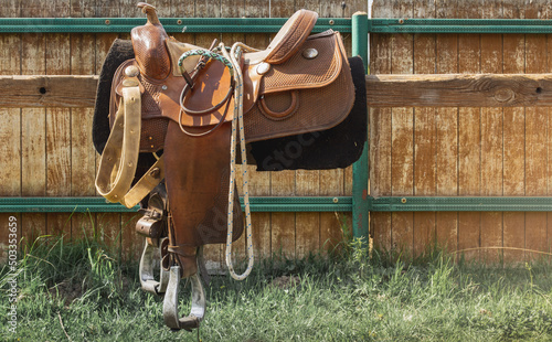 Brown leather saddle and equipment for riding. West style. Decorative bridle, ropes, stirrup. Wooden paddock fence background