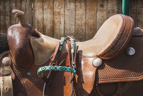 Brown leather saddle and equipment for riding. West style. Decorative bridle, ropes, stirrup. Wooden paddock fence background