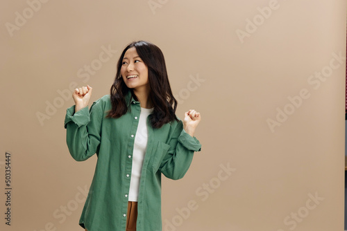 Canvas Print Overjoyed cute Asian student young woman in khaki green shirt raise fists up say Yeah posing isolated on over beige pastel studio background