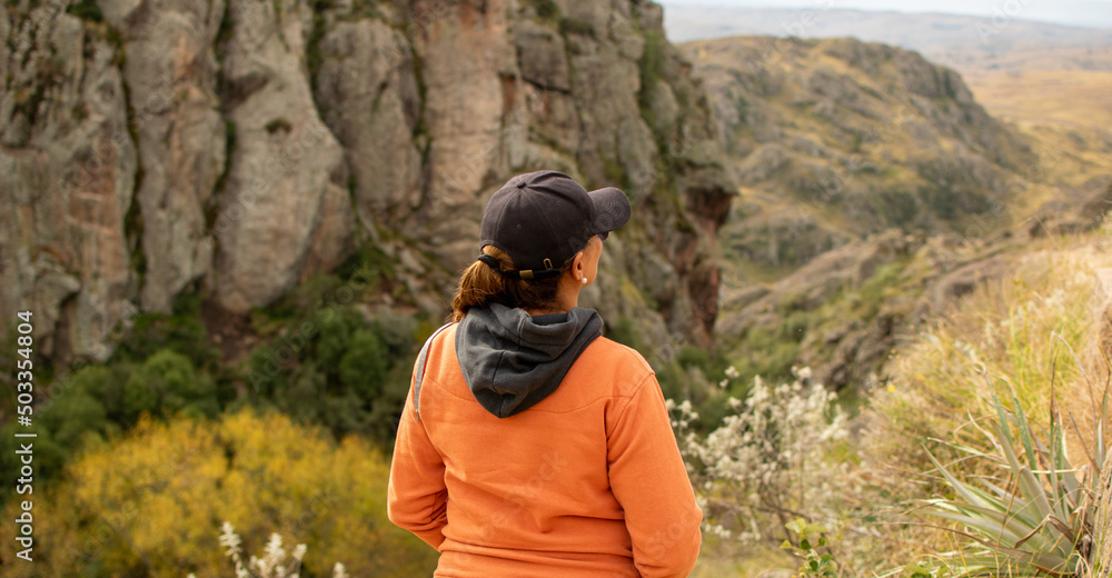 Naklejka premium Portrait of latin woman dressed in orange with a cap and tail turned back having fun during the day of trekking in the mountain forest - Focus from the back