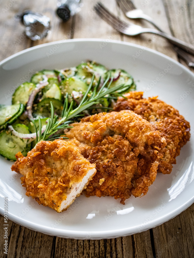 Breaded fried pork chop with sliced cucumbers and rosemary on wooden table
