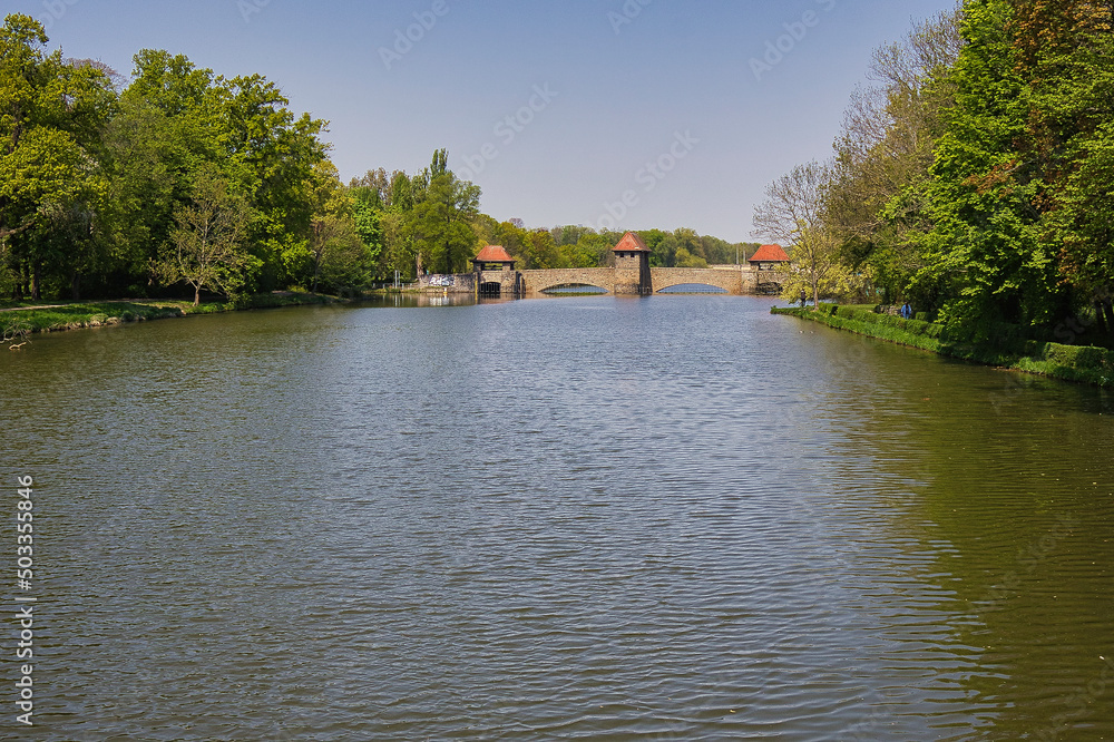 Fototapeta premium Blick von der Klingerbrücke über das Elsterbecken auf das historische Palmengartenwehr im Frühling mit grüner Uferböschung, Leipzig, Sachsen, Deutschland