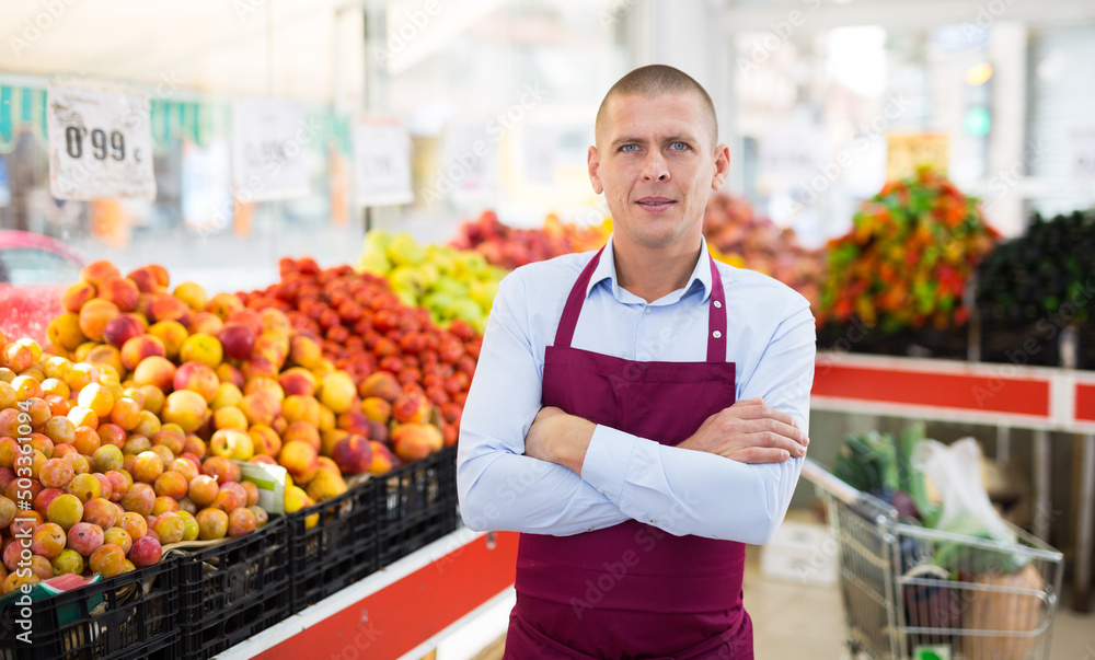 Male worker in apron standing in salesroom of greengrocer and looking in camera.