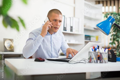 Portrait of focused manager sitting at office table, having phone conversation