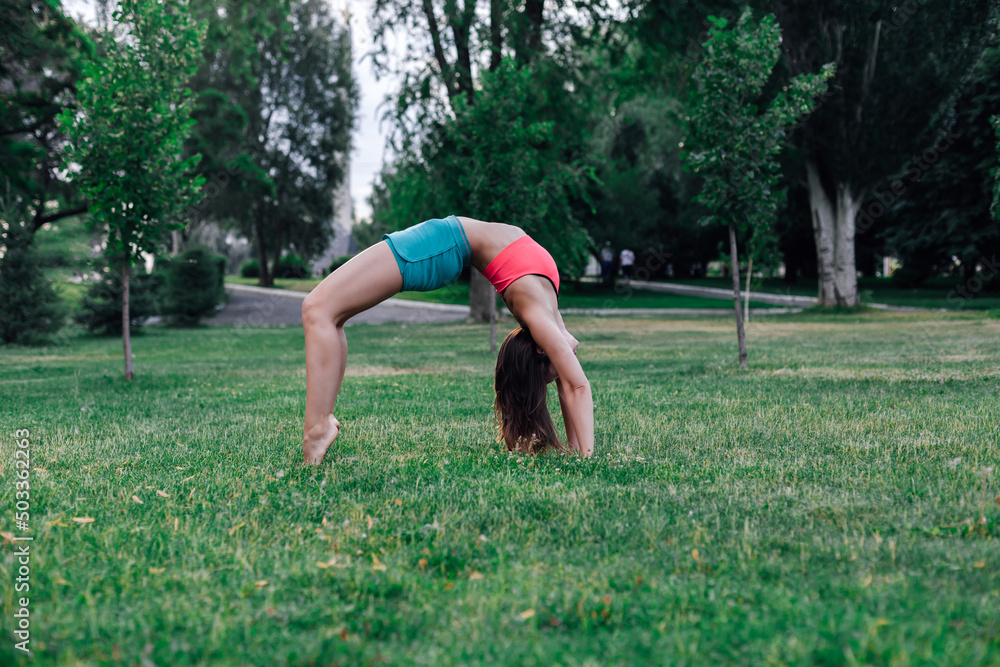 Beautiful young woman trains in nature. Sporty brunette in pink top and blue shorts, doing yoga, standing in bridge pose, full-length on green grass. Beautiful young woman trains in nature. Sporty brunette in pink top and blue shorts, doing yoga, standing in bridge pose, full-length on green grass.