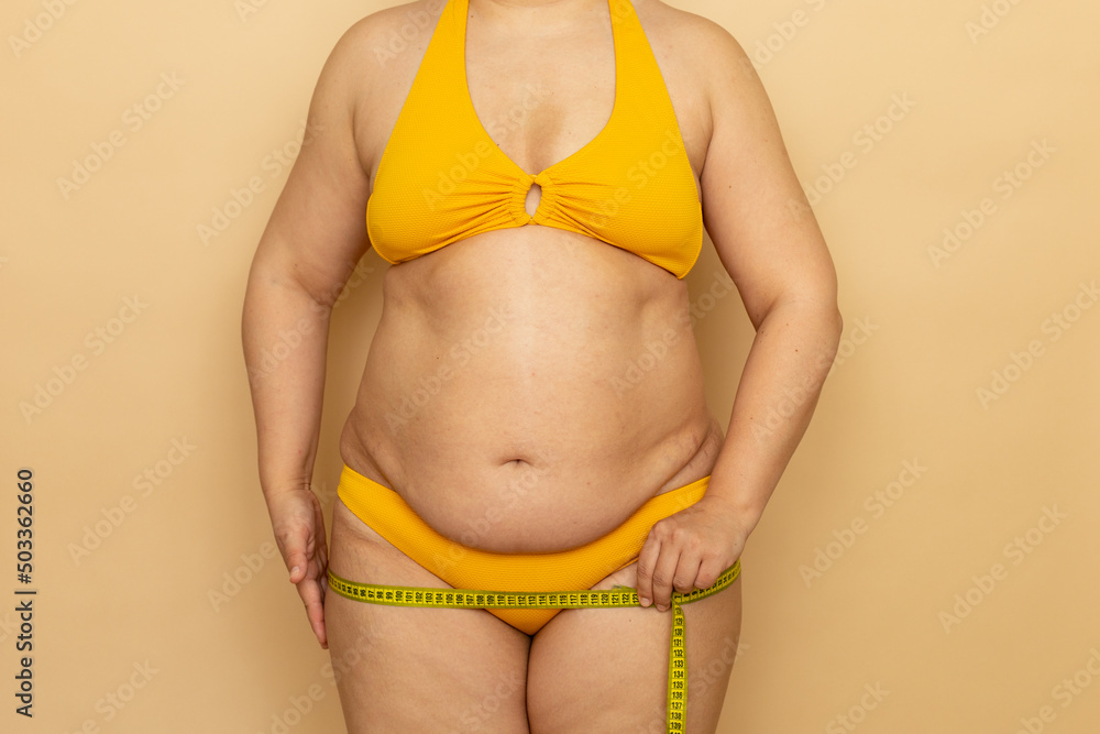 Overweight woman in yellow swimsuit measure hips circumference with measuring tape, beige background. Slimming and body control, struggle with obesity. Plus size people and body positive concept. Overweight woman in yellow swimsuit measure hips circumference with measuring tape, beige background. Slimming and body control, struggle with obesity. Plus size people and body positive concept.