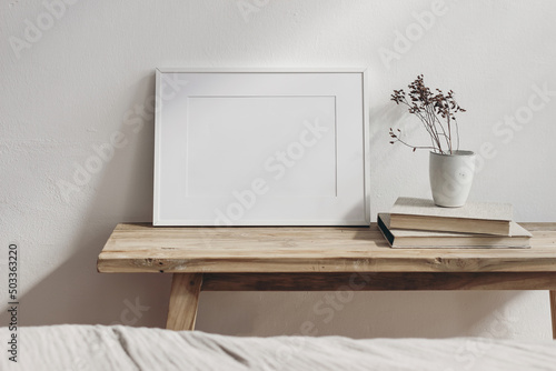 Portrait white frame mockup on vintage wooden bench, table. Little ceramic vase with dry hypericum plant on pile of books. Blurred beige linen bed in front. Modern Scandinavian interior.