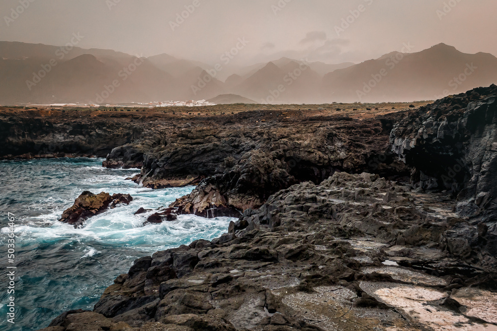 Rocky shore of Tenerife. Black coast with mountains on background. Rocky shore of Tenerife. Black coast with mountains on background.