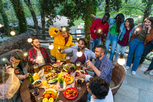 Young people dining and singing with phone flashlight on – group friend having fun drinking wine in dinner party – dinning and drinking on terrace restaurant – happy people eating food together