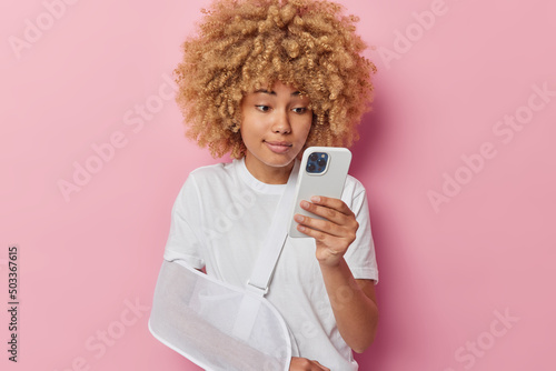 Photo of young female model has injured arm in sling uses mobile phone for scrolling social networks finds advice for treatment in internet isolaed over pink background. Insurance accident concept photo