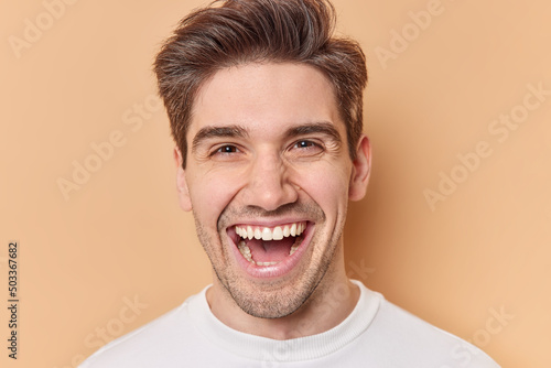 Portrait of handsome cheerful man laughs gladfully smiles toothily being in good mood expresses positive emotions wears casual white jumper isolated over brown background. Happiness concept. © wayhome.studio 