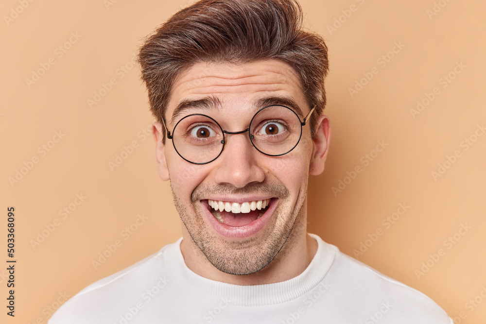 Portrait of happy surprised adult man reacts on amazing news wears round transparent eyeglasses smiles broadly dressed in casual wear isolated over brown background. Human reactions concept. Portrait of happy surprised adult man reacts on amazing news wears round transparent eyeglasses smiles broadly dressed in casual wear isolated over brown background. Human reactions concept.