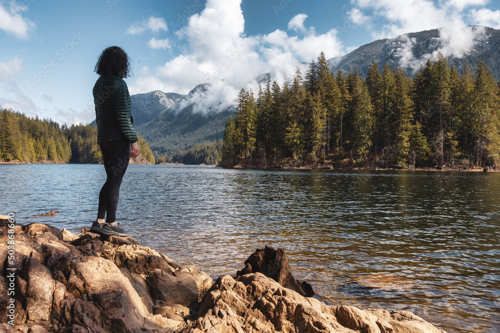 Adventurous Caucasian Woman on the rocks by the water in Canadian Nature Landscape. Buntzen Lake, Anmore, Vancouver, BC, Canada. Adventure Travel Concept