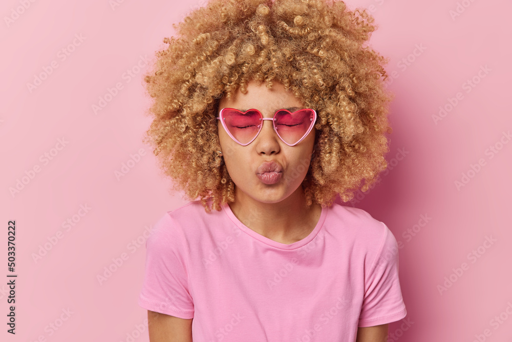 Romantic beautiful woman with curly hair keeps lips folded blows air kiss at camera closes eyes wears heart sunglasses and casual pink t shirt in one tone with background. Facial expressions concept