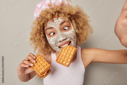Funny curly haired woman eats delicious waffles makes grimace crosses eyes wears bathhat and casual t shirt poses for selfie applies beauty mask for skin treatment isolated over grey background