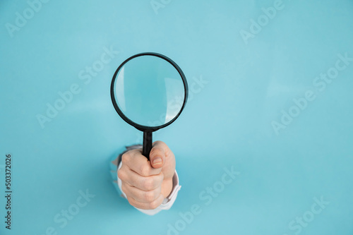 A woman's hand sticking out of a hole in a paper blue background holds a magnifying loupe. Copy space. 