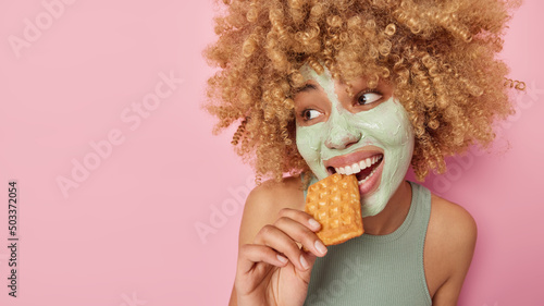 Pretty positive woman with curly bushy hair bites delicious waffle applies beauty mask on face for skin treatment undergoes anti aging procedures wears casual t shirt isolated over pink wall