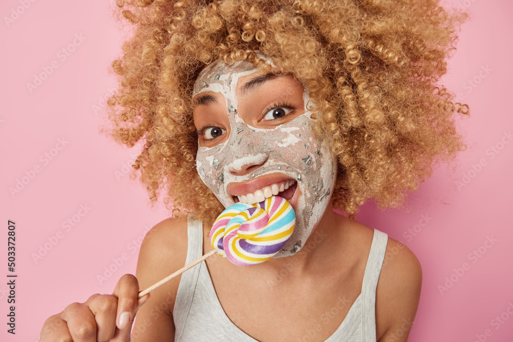Close up portrait of cheerful curly haired woman bites caramel candy applies beauty mask on face for skin treatment wears casual t shirt isolated over pink background. Unhealthy food concept