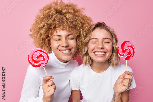 Portrait of cheerful women have happy expressions smile broadly hold lollipops on sticks have sweet tooth enjoy eating sweet candies dressed in casual white t shirts isolated over pink background