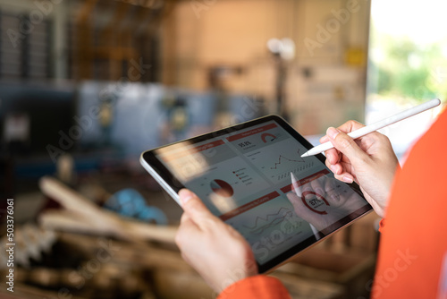 Action of a working is using tablet to review factory report with blurred background of warehouse. Business management and technology concept photo. Close-up and selective focus at human's hand part.