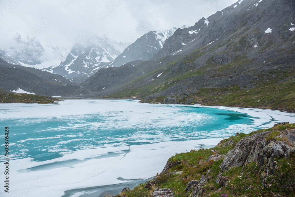 Atmospheric mountain landscape with frozen alpine lake during snowfall. Awesome cold scenery with snowfall in high mountain valley with icy mountain lake on background of snow mountains in low clouds. Atmospheric mountain landscape with frozen alpine lake during snowfall. Awesome cold scenery with snowfall in high mountain valley with icy mountain lake on background of snow mountains in low clouds.