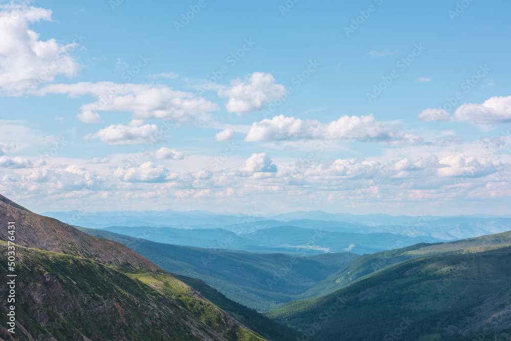 Scenic aerial landscape with green mountain vastness under cumulus clouds in blue sky in changeable weather. Colorful awesome top view to forest valley among sunlit hills and mountains on horizon.