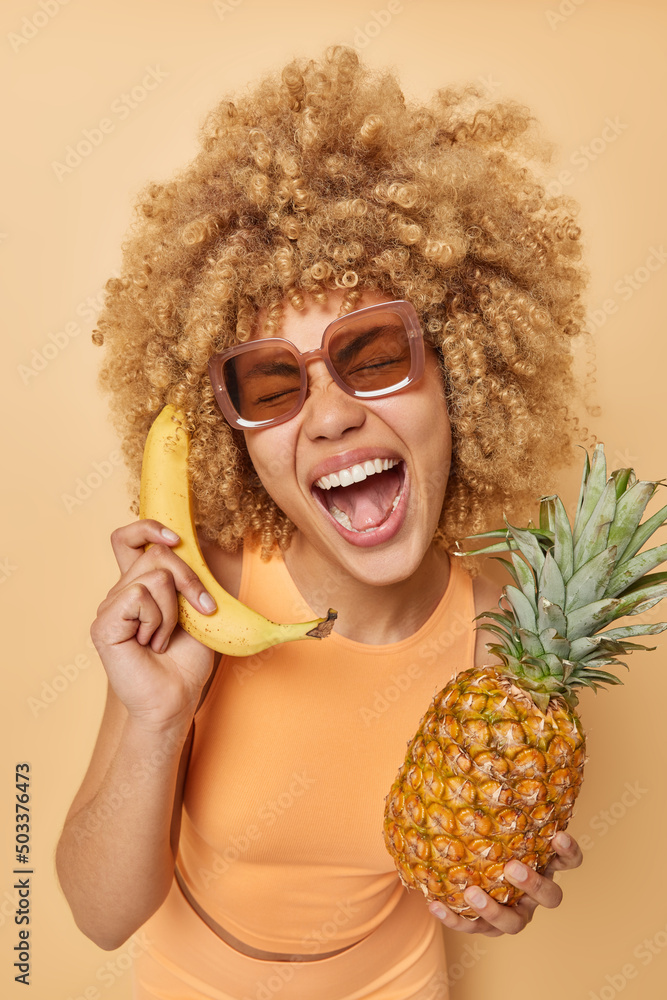 Cheerful woman has playful expression holds fresh pineapple and banana near  ear pretends having telephone talk keeps mouth opened wears sunglasses  sportswear isolated over beige studio background Stock Photo | Adobe Stock