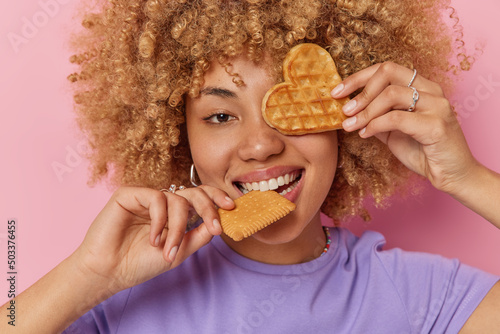 Headshot of carefree curly teenage girl bites cookies covers eye with appetizing waffle has sweet tooth enjoys eating delicious food wears casual purple t shirt isolated over pink background