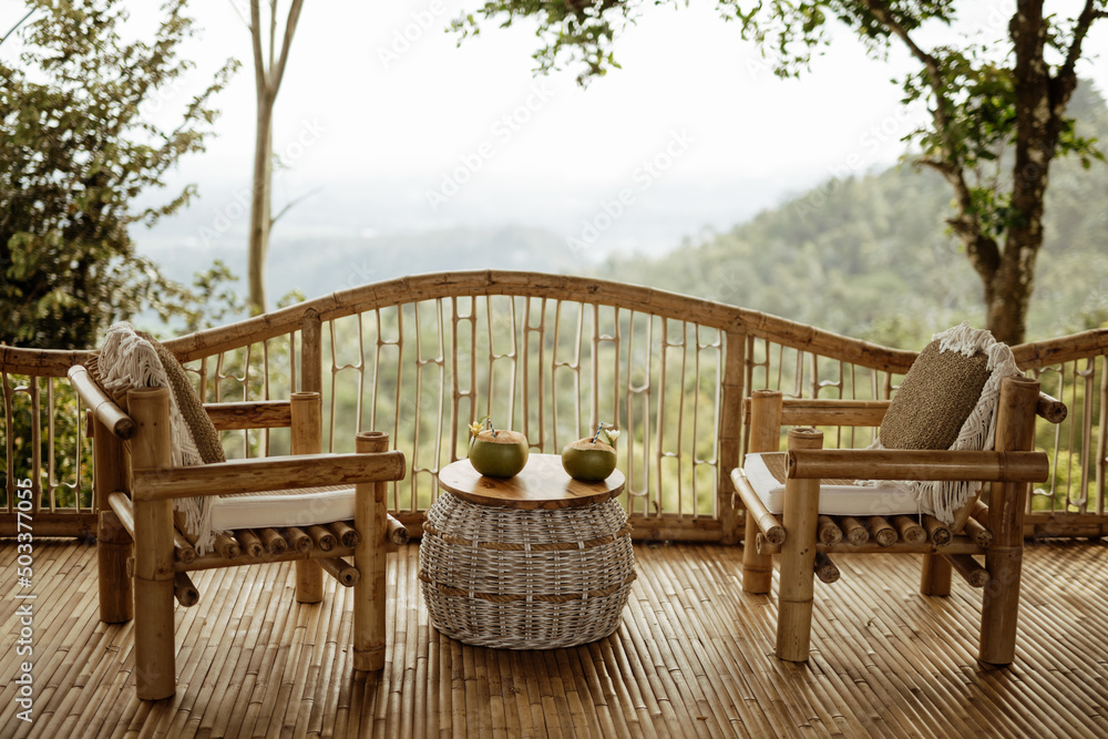 Obraz premium Couple chairs on balcony of countryside bamboo terrace house with mountain view background in the morning. Two coconuts on the table, couple vacation concept