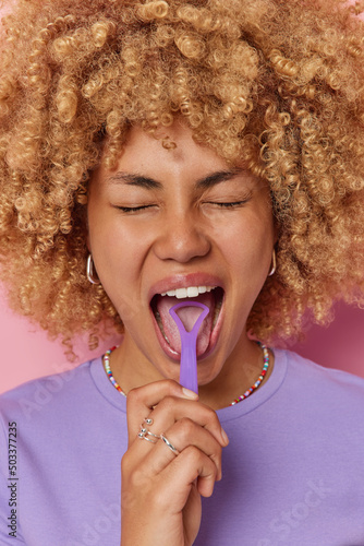 Vertical shot of curly haired feamle model uses tongue scraper takes care of oral hygiene dressed in purple t shirt cleans mouth poses indoors. Young beautiful woman uses tongue cleaner regularly