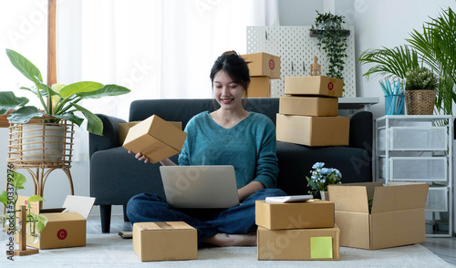 Happy young asian woman startup small business freelance holding parcel box and computer laptop and sitting on floor, Online marketing packing box delivery concept © wichayada