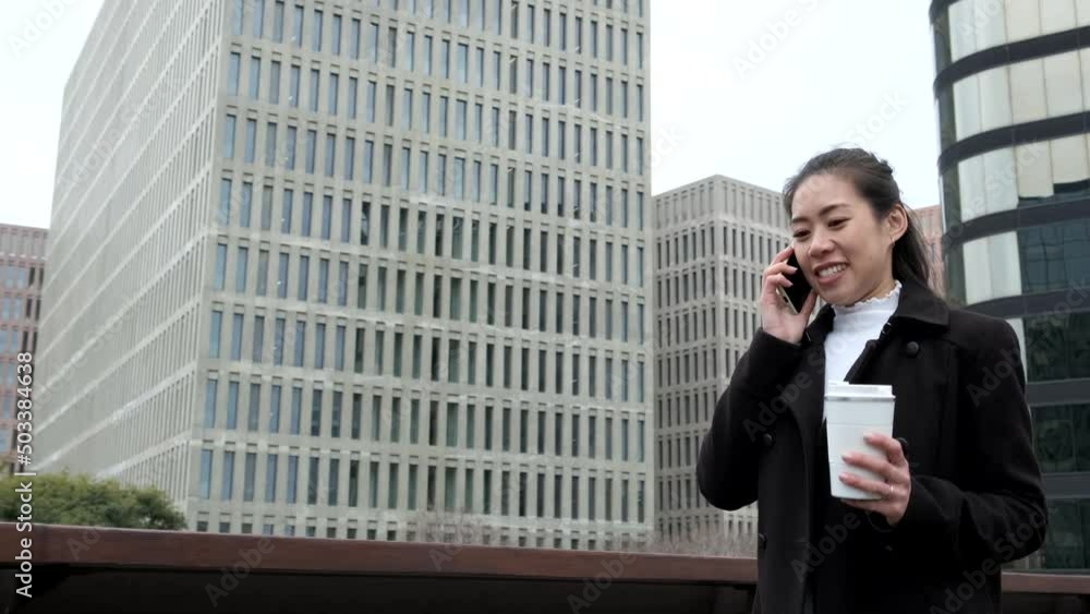 From below tracking shot of happy Asian female entrepreneur in black coat with coffee to go. She is smiling and having smartphone conversation while walking against skyscrapers in downtown From below tracking shot of happy Asian female entrepreneur in black coat with coffee to go. She is smiling and having smartphone conversation while walking against skyscrapers in downtown