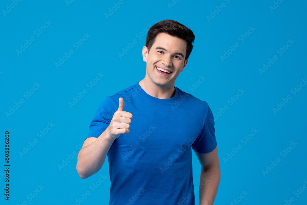 Fototapeta premium Smiling handsome young Caucasian man giving thumbs up in light blue isolated background