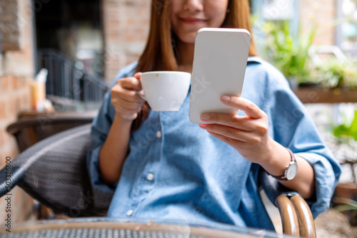 Closeup image of a young woman holding and using mobile phone while drinking coffee in the outdoors cafe