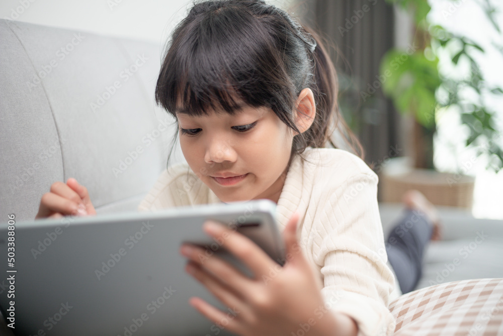 Little girl using tablet playing game on the internet, Kid sitting on sofa watching or talking with a friend online, Child relaxing in living room in the morning, Children with New Technology concept Little girl using tablet playing game on the internet, Kid sitting on sofa watching or talking with a friend online, Child relaxing in living room in the morning, Children with New Technology concept
